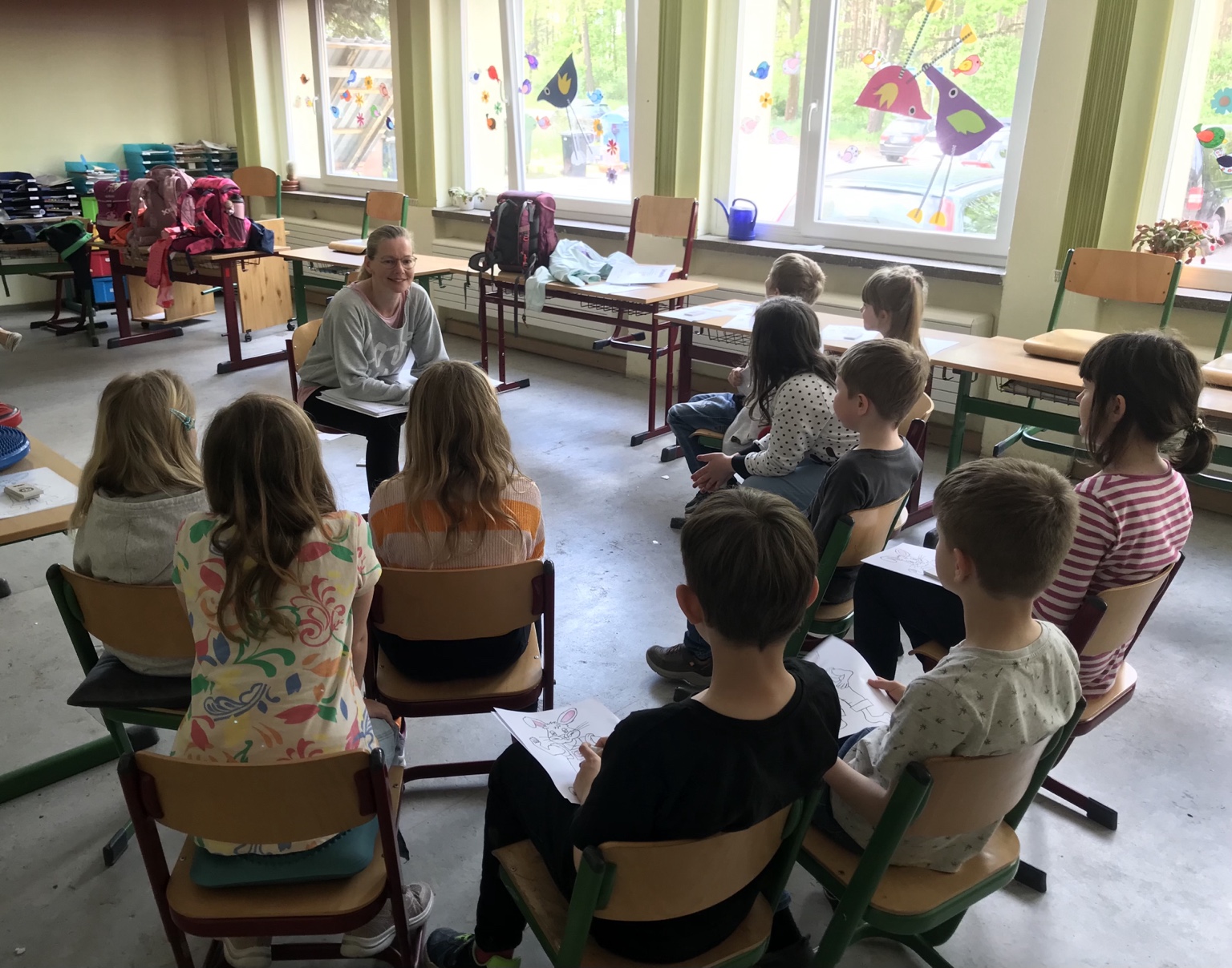 Nadine im Gespräch mit Kindern im Sitzkreis eines Klassenzimmers; die Kinder halten Pauli-Ausmalblätter.