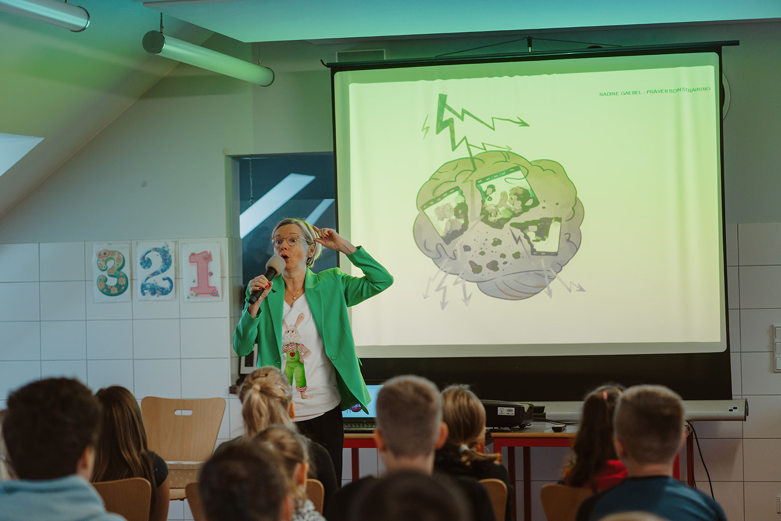 Nadine spricht mit Mikrofon vor Kindern; auf der Leinwand ist eine Illustration des Gehirns zu sehen.
